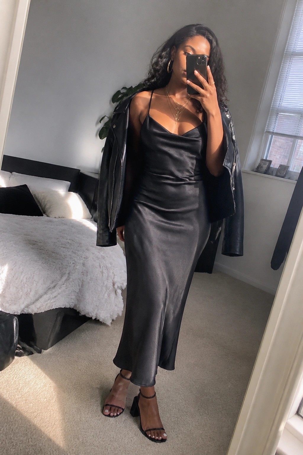 Woman in black satin slip dress and leather jacket taking bedroom mirror selfie.