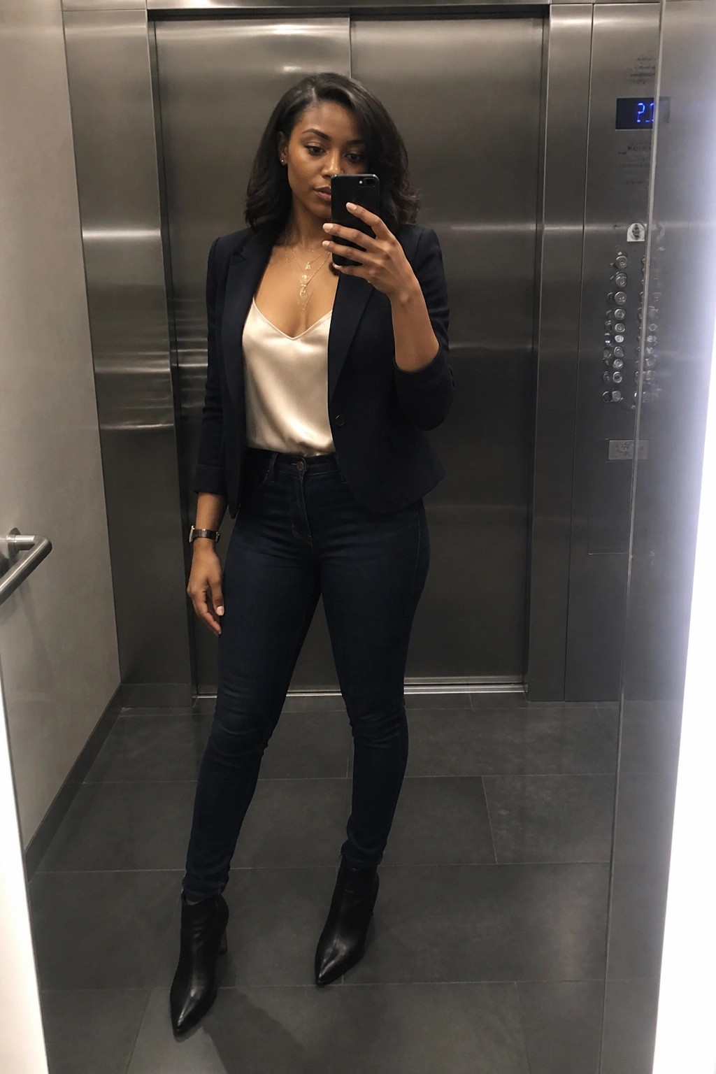 Woman standing in elevator wearing navy blazer over cream satin camisole paired with dark skinny jeans black pointed ankle boots and layered necklaces while holding a phone for a mirror selfie.