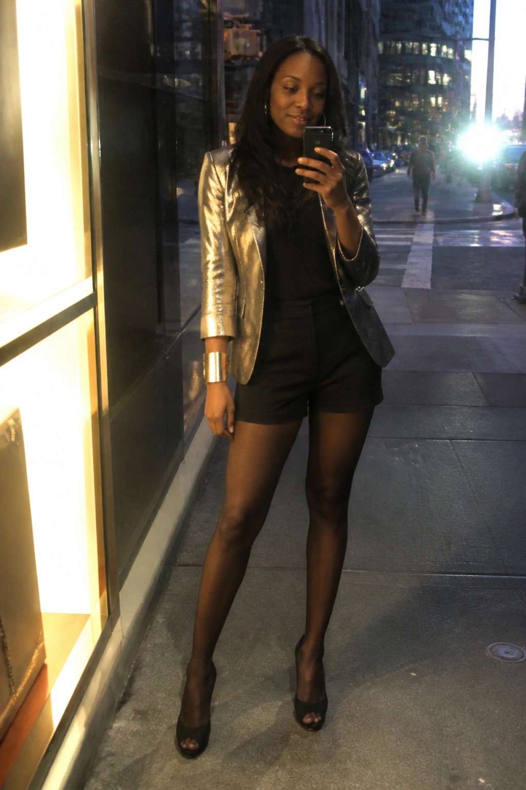 Woman wearing a shiny gold metallic blazer over a black top and black shorts with sheer black tights black peep toe heels and a wide gold cuff bracelet while holding a phone on a city sidewalk at night.