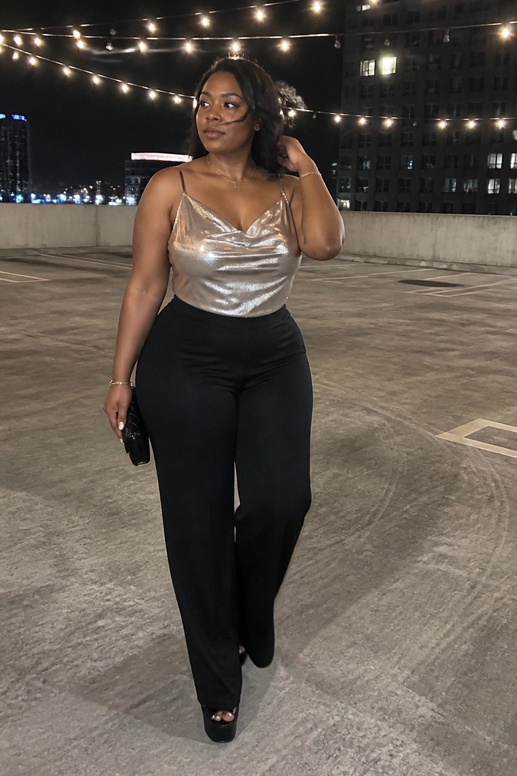 Curvy woman in metallic silver camisole and black wide-leg pants on rooftop at night