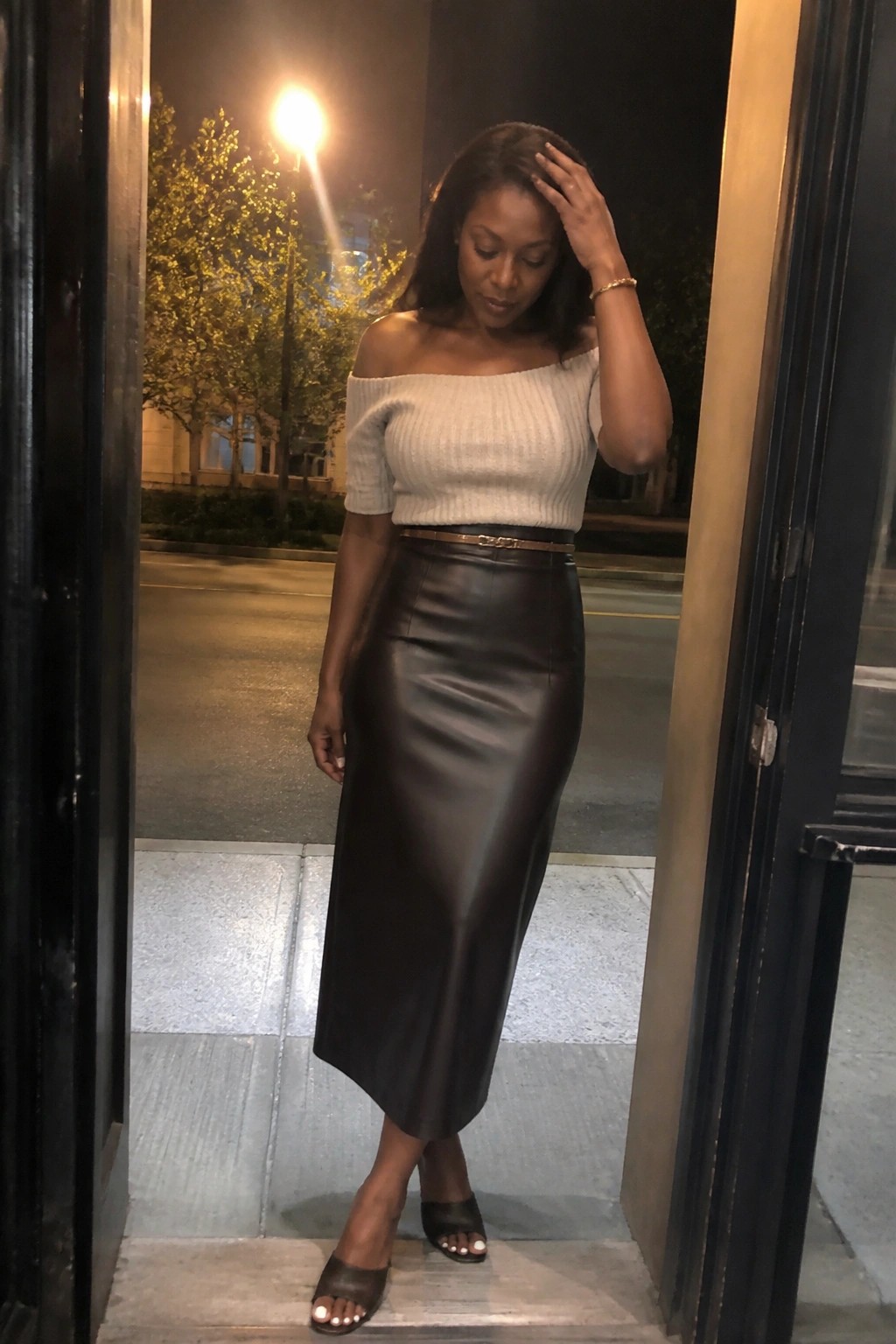 Woman in beige off-shoulder sweater and brown leather midi skirt at night doorway.