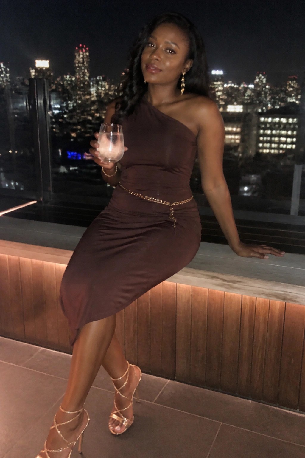 Woman in brown one-shoulder dress holding cocktail on balcony overlooking city lights at night