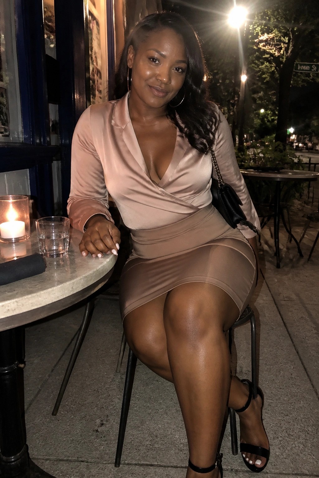 Woman in beige satin dress seated at outdoor cafe table with lit candle at night