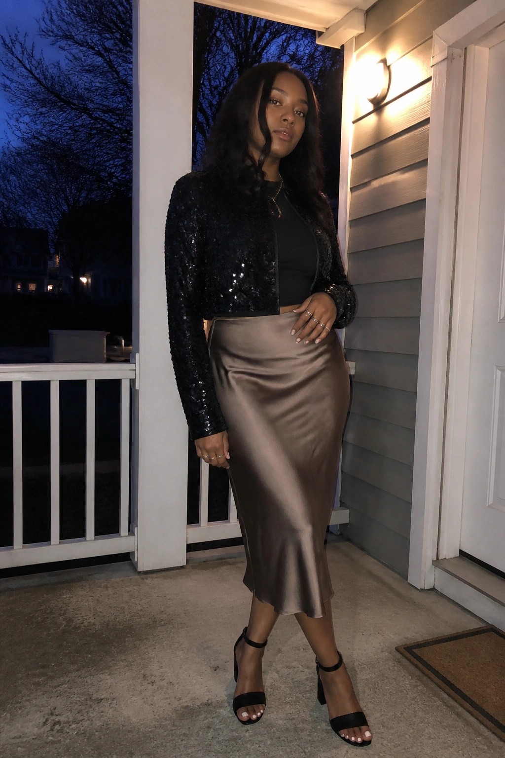 Woman in black sequined jacket and taupe satin skirt on porch at night.