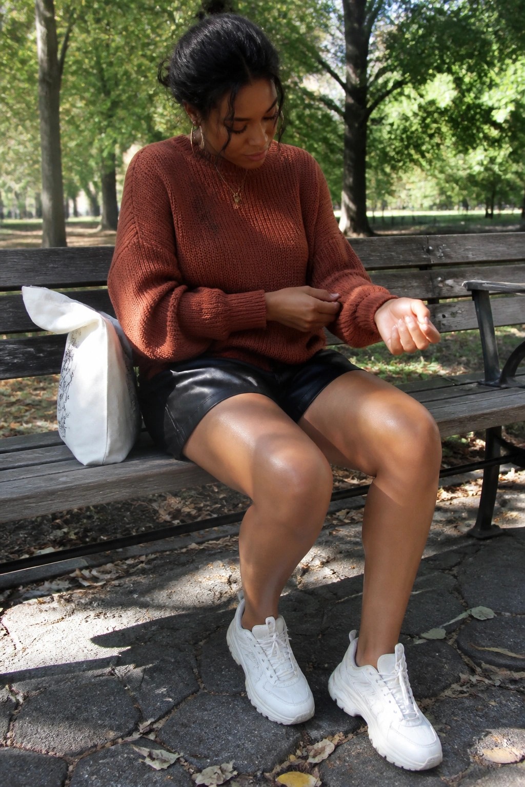 Woman in rust sweater and black shorts sits on park bench with white tote bag.