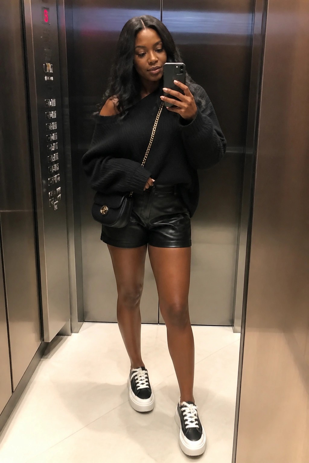 Woman taking mirror selfie in elevator wearing black off-shoulder sweater and leather shorts