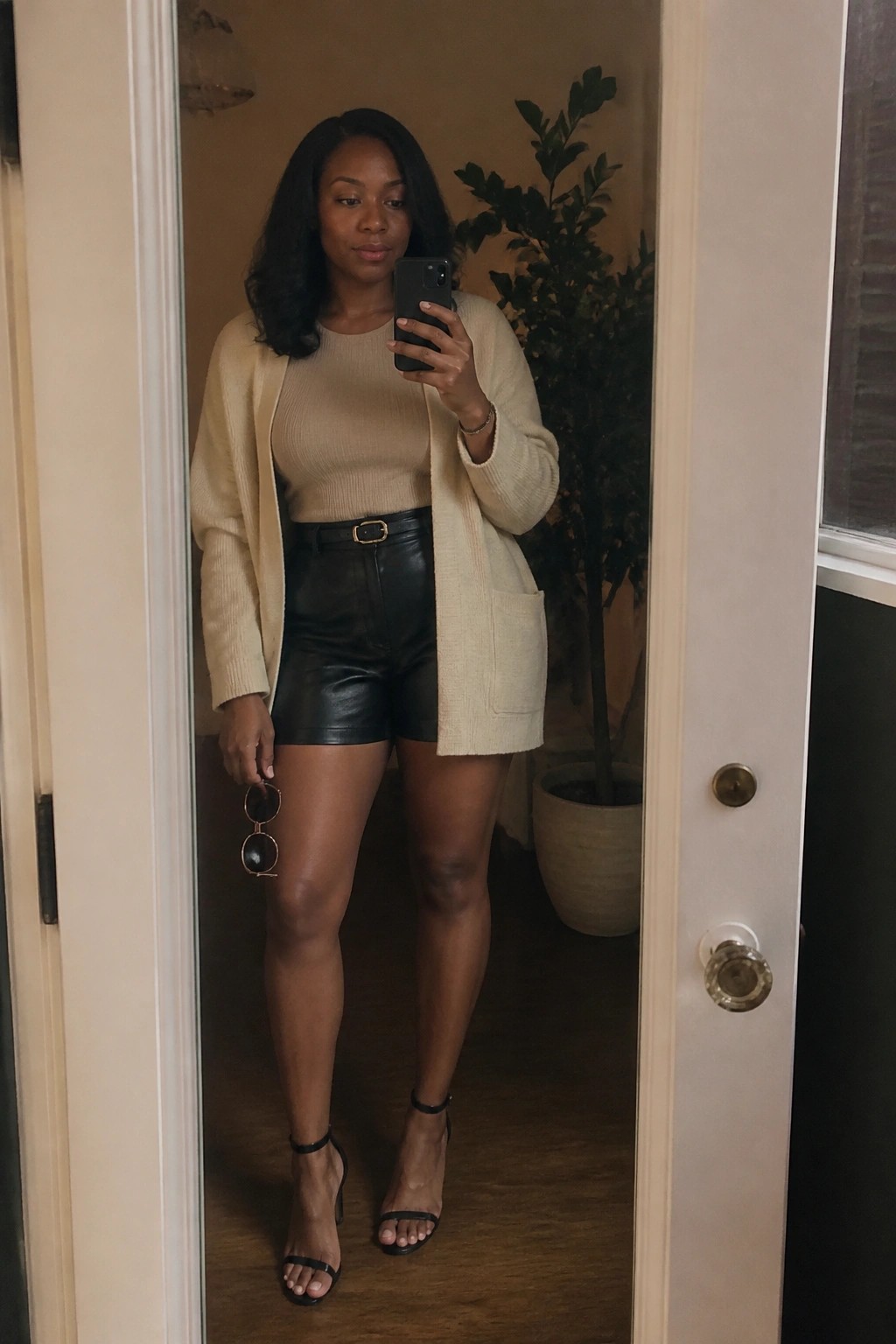 Woman wearing an oversized beige knit cardigan over a beige ribbed top with black leather shorts belted at the waist and black strappy heels while holding a phone and sunglasses in a doorway.