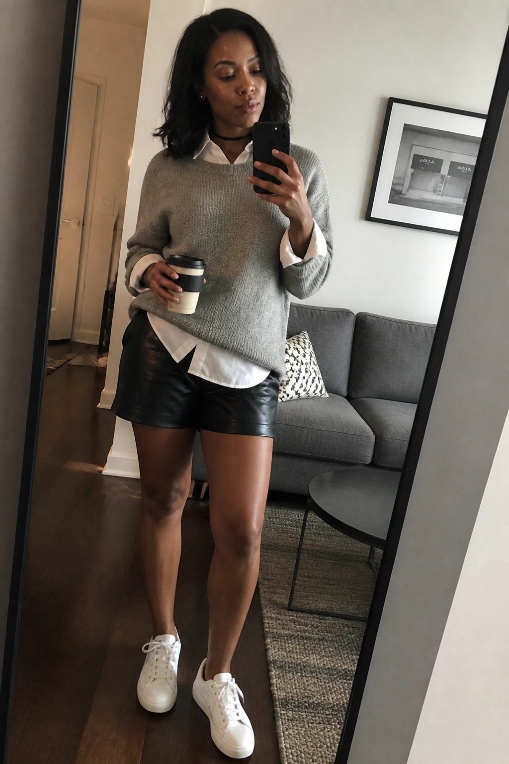 Woman in gray sweater over white shirt with black leather shorts holding coffee and phone