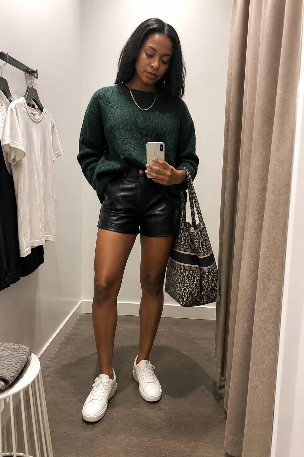 Woman in green sweater and black leather shorts checks phone in fitting room
