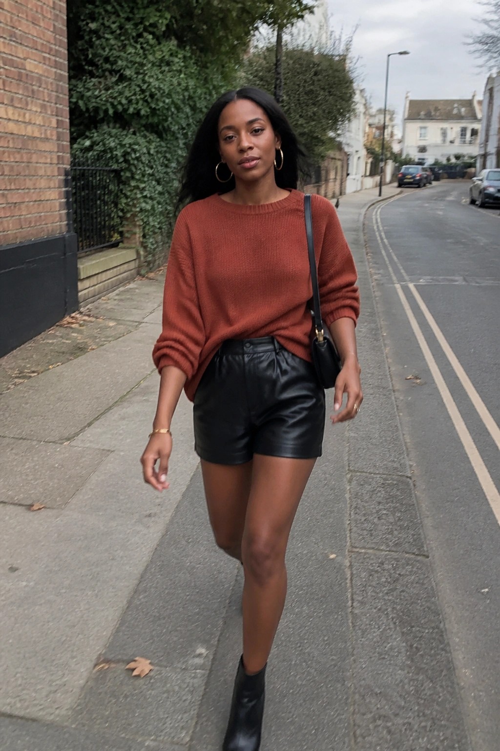 Woman in rust sweater and black leather shorts walks on urban sidewalk