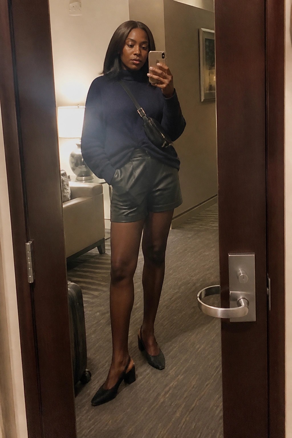 Woman in navy turtleneck and black leather shorts taking mirror selfie in hotel