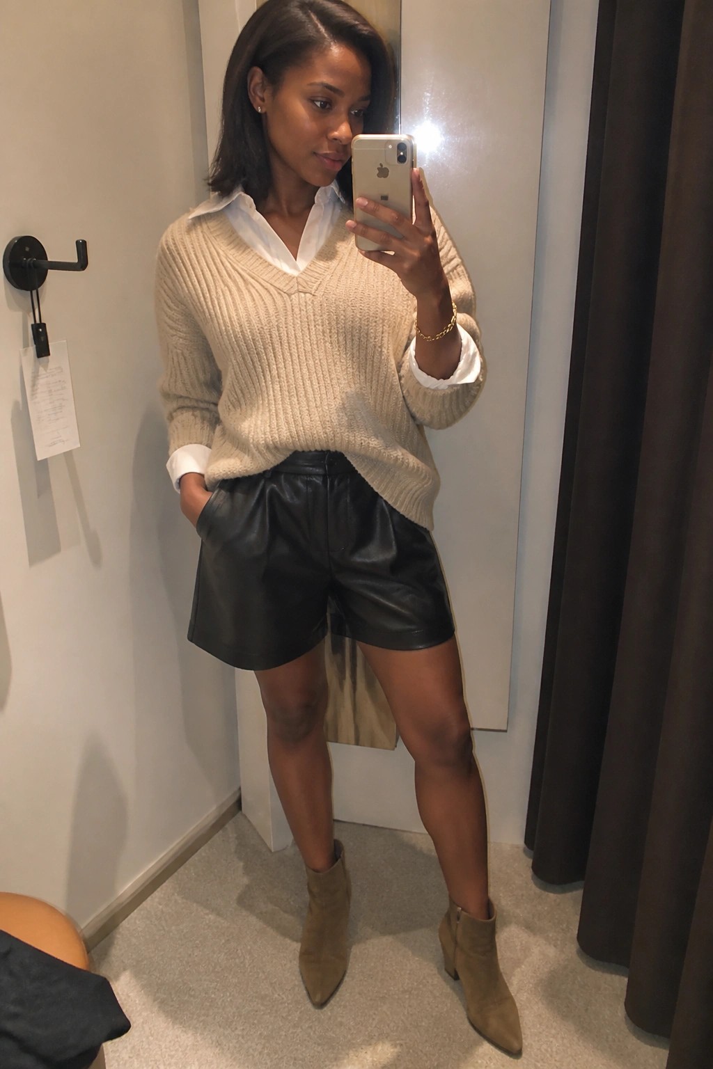 Woman in beige knit sweater, white shirt, black leather shorts, and suede boots taking mirror selfie