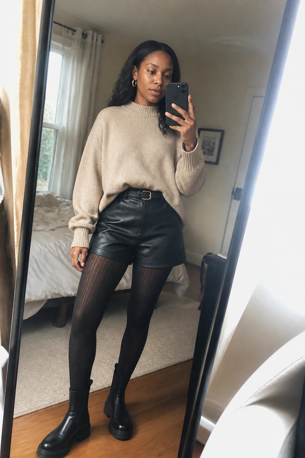Woman in beige sweater and black leather shorts taking mirror selfie in bedroom.