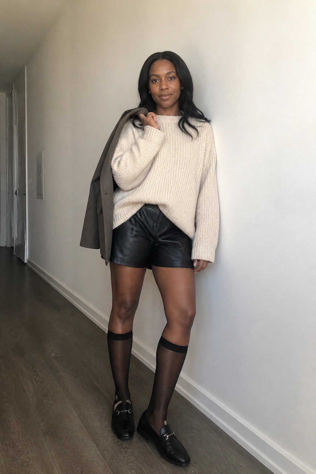 Woman in cream knit sweater, black leather shorts, sheer socks, and gray blazer over shoulder