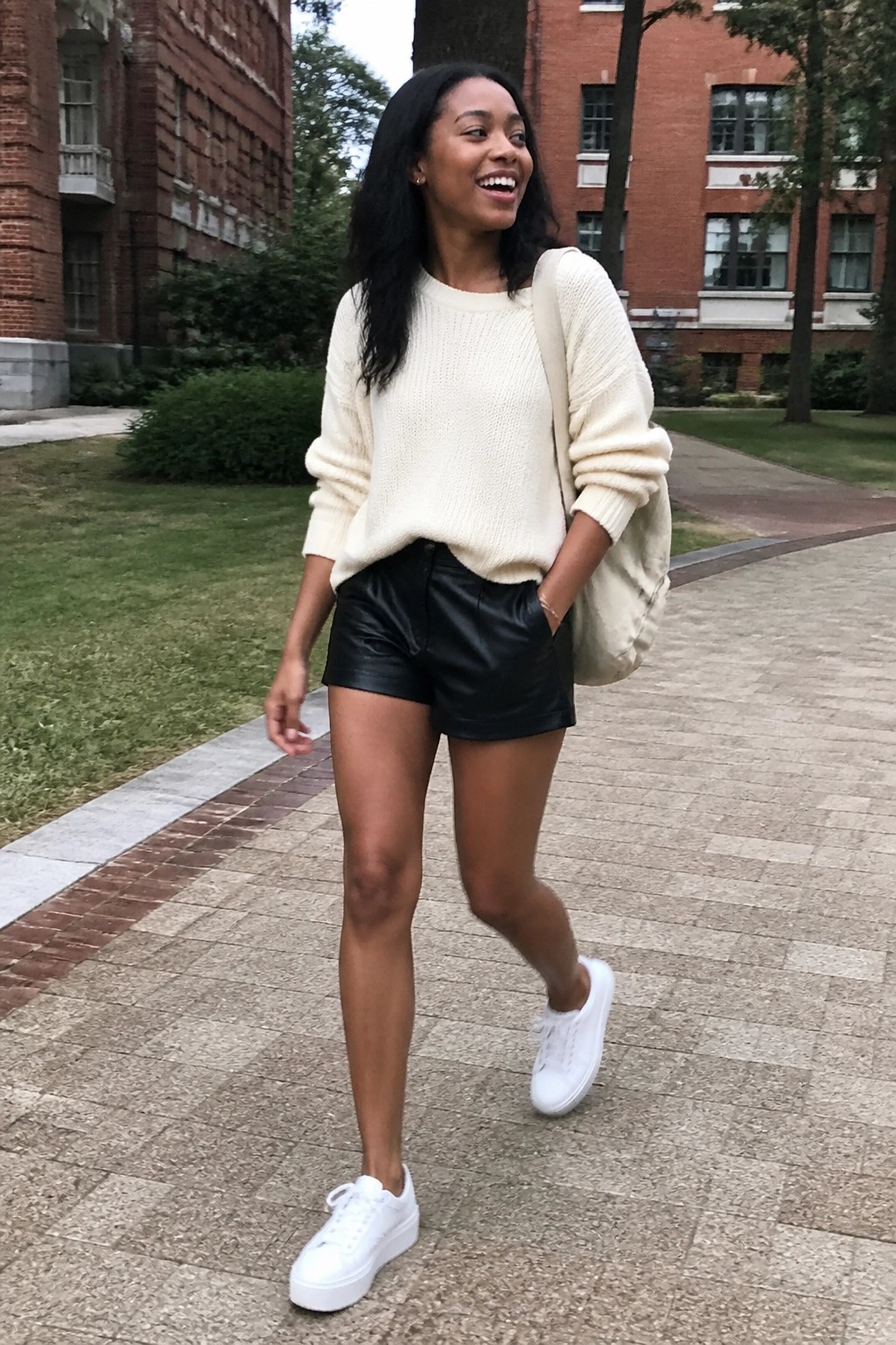 Woman in cream sweater and black leather shorts walks on brick path with backpack