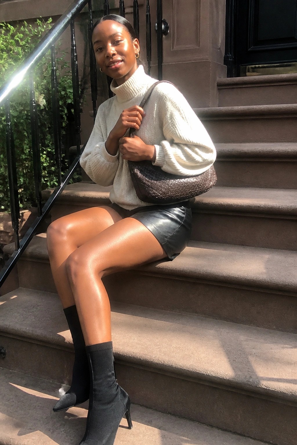 Black woman seated on outdoor steps in oversized cream turtleneck sweater tucked slightly over black leather shorts with black pointed toe heeled boots and brown textured handbag held in hands