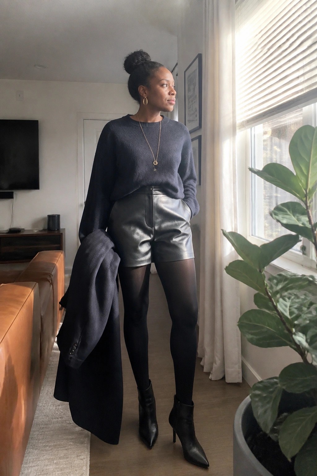 Woman in navy sweater and metallic shorts holding coat by sunlit window