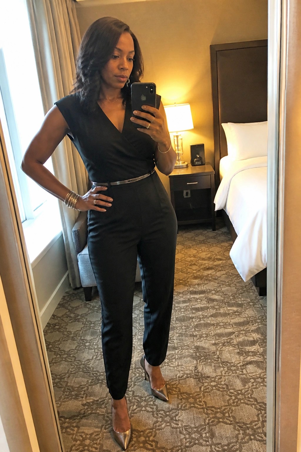 Woman in black jumpsuit and silver heels taking mirror selfie in hotel room
