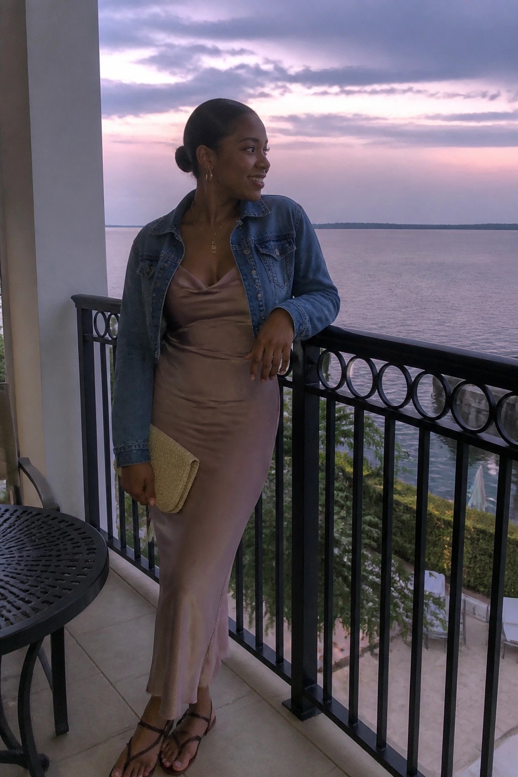 Woman in denim jacket over satin dress on balcony overlooking water at dusk