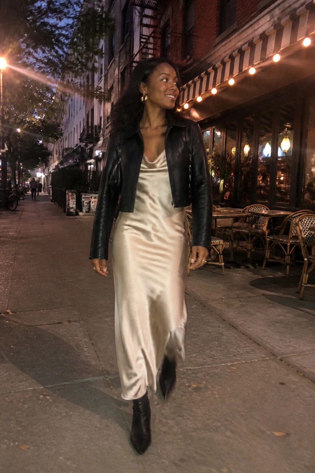 Black woman wearing a cropped black leather jacket over a champagne satin slip dress paired with black ankle boots and gold jewelry.
