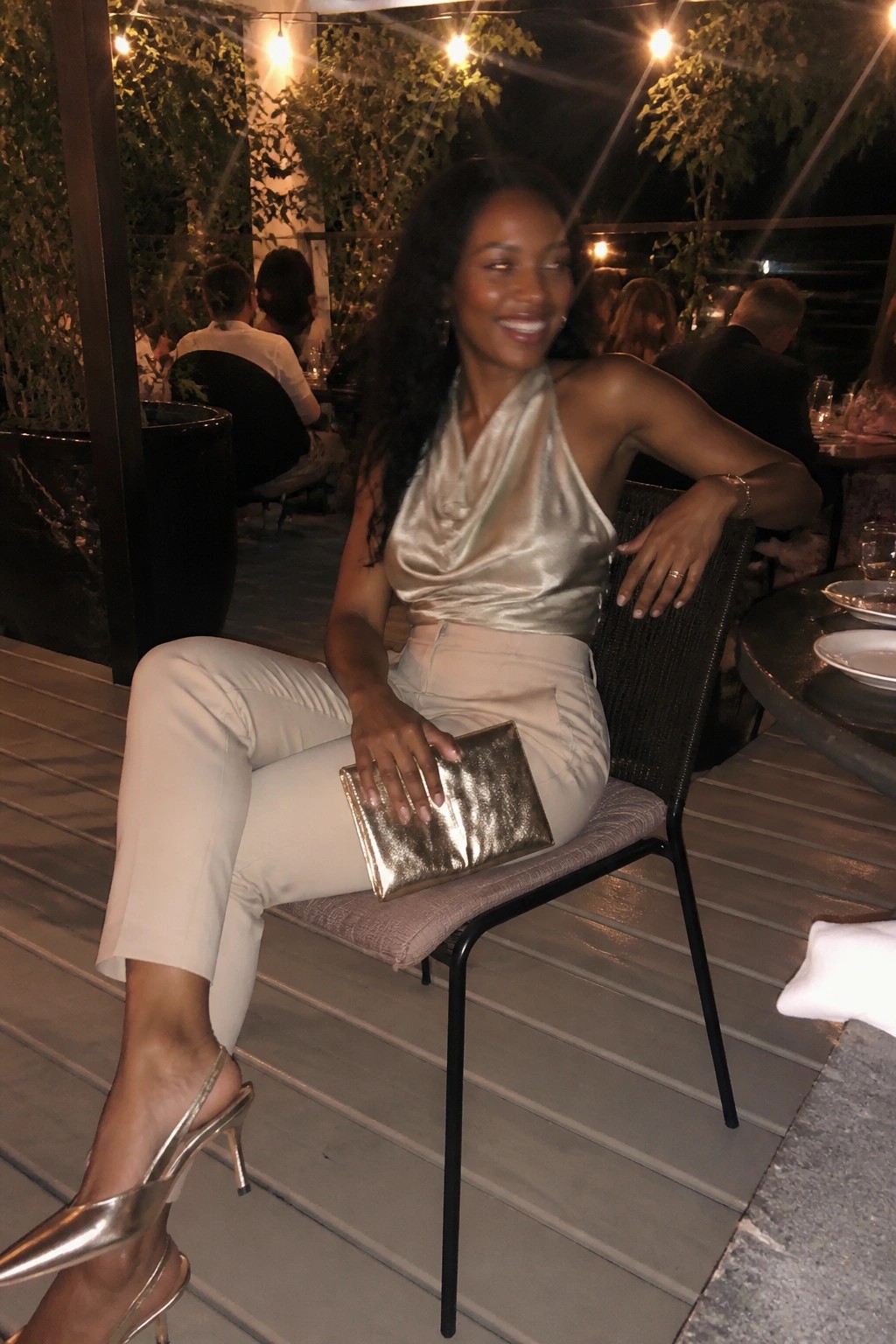 Woman in gold satin halter top and beige pants holding metallic clutch at night restaurant
