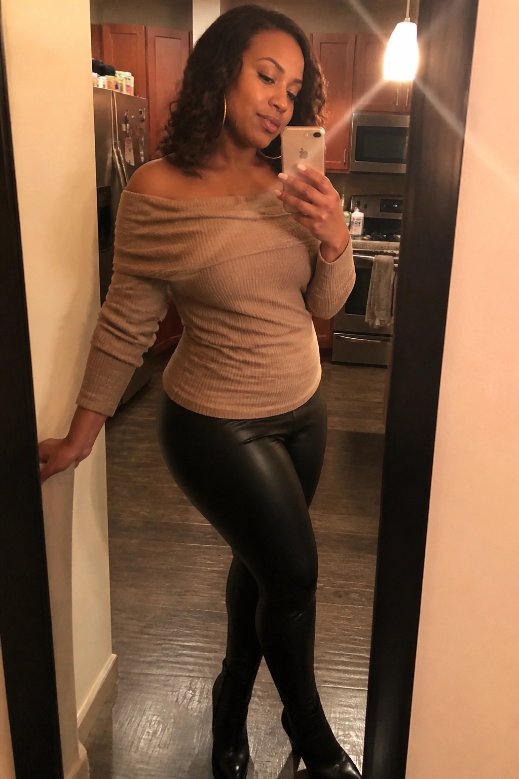 Woman in beige off-shoulder sweater and black leggings taking mirror selfie in kitchen