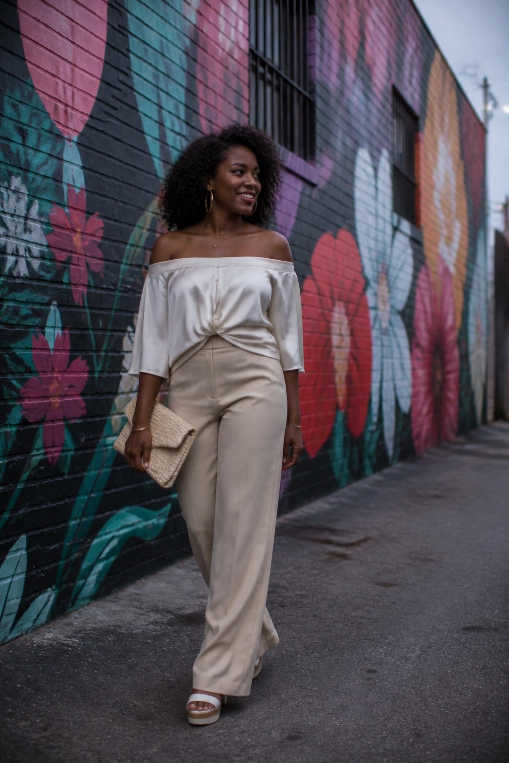 Woman with curly hair in off-shoulder top and beige pants before floral mural