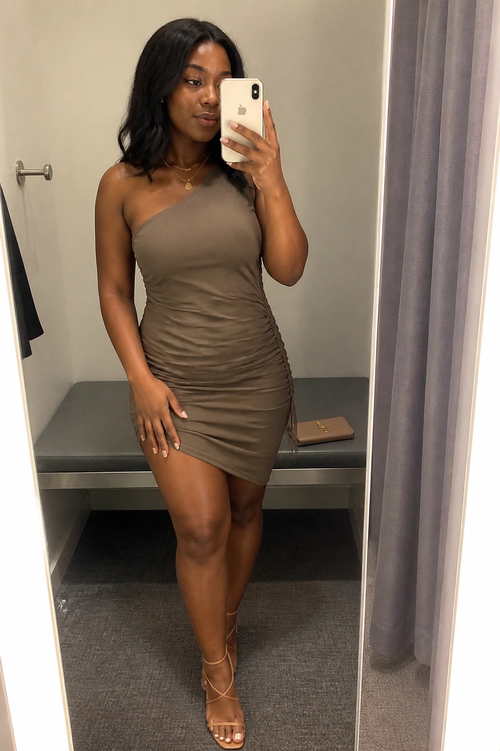 Woman in taupe one-shoulder ruched mini dress taking mirror selfie in fitting room