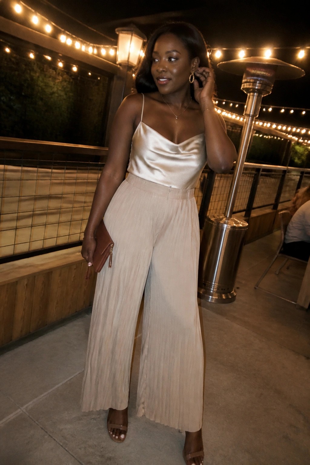 Woman in beige satin camisole and pleated pants posing on string-lit patio at night.