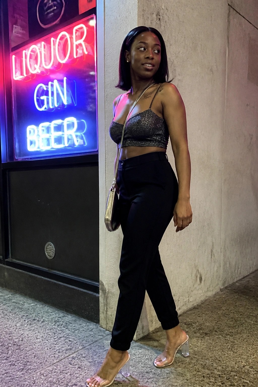 Woman in sequined crop top and black pants beside red-blue neon liquor sign.