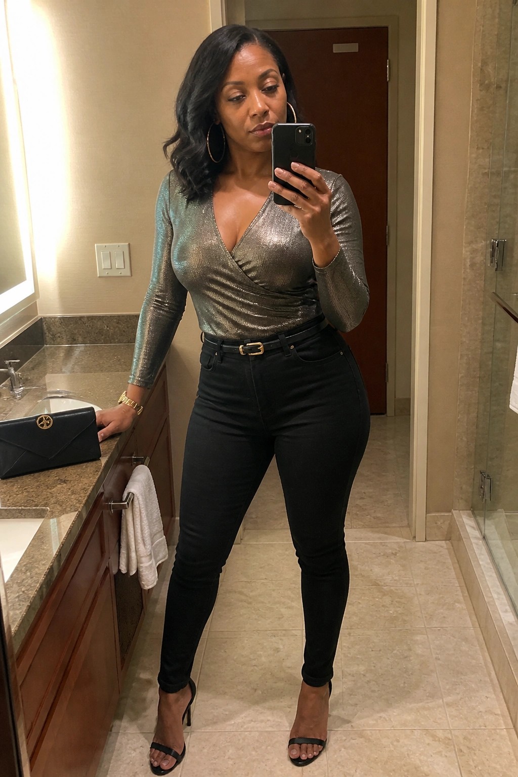 Woman in metallic top and black jeans taking mirror selfie in bathroom.