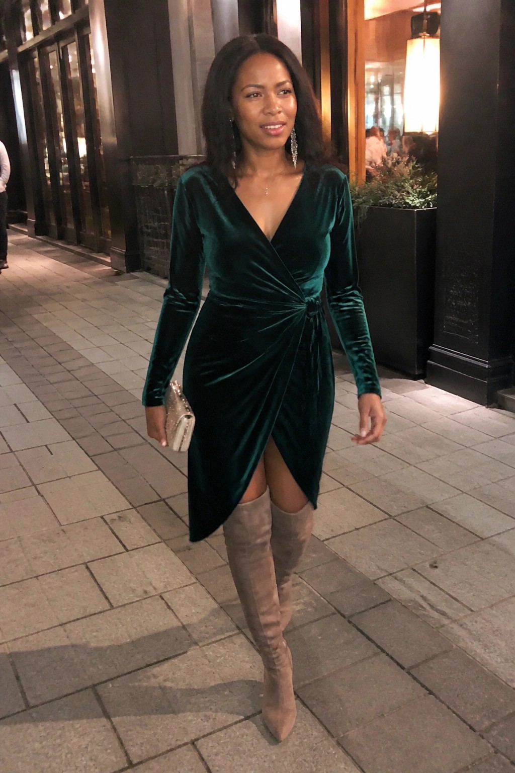 A Black woman in a dark green velvet wrap dress with long sleeves, a front twist detail, and a thigh slit, paired with beige over-the-knee suede boots while holding a small embellished clutch.
