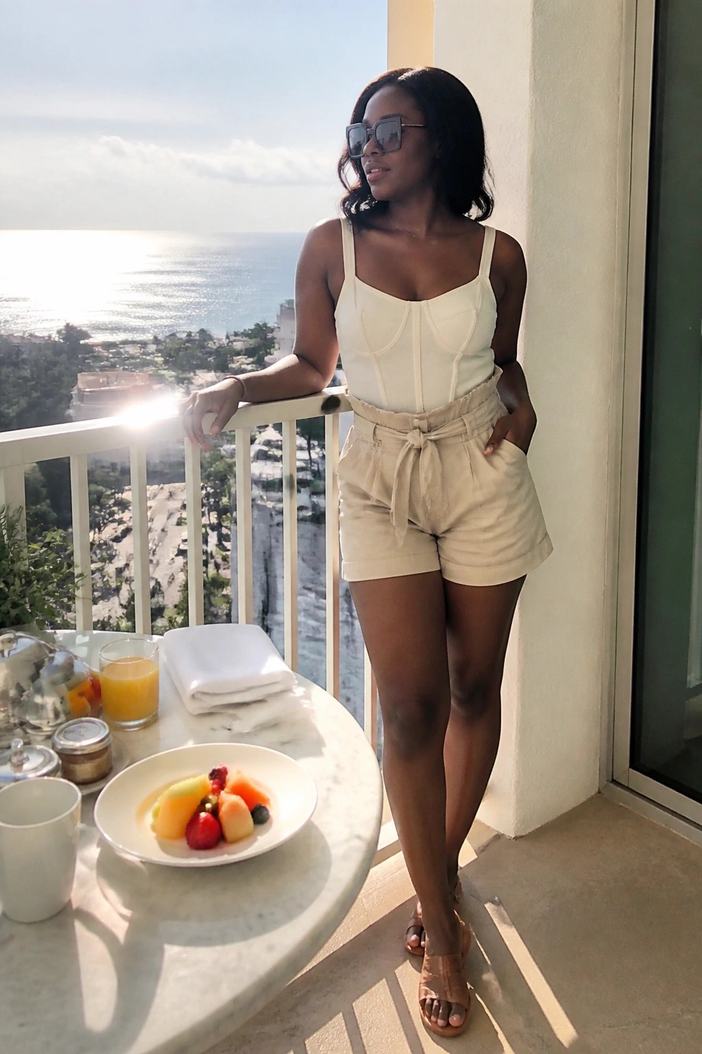 Woman in white corset top and beige shorts on sunny ocean-view balcony with breakfast table