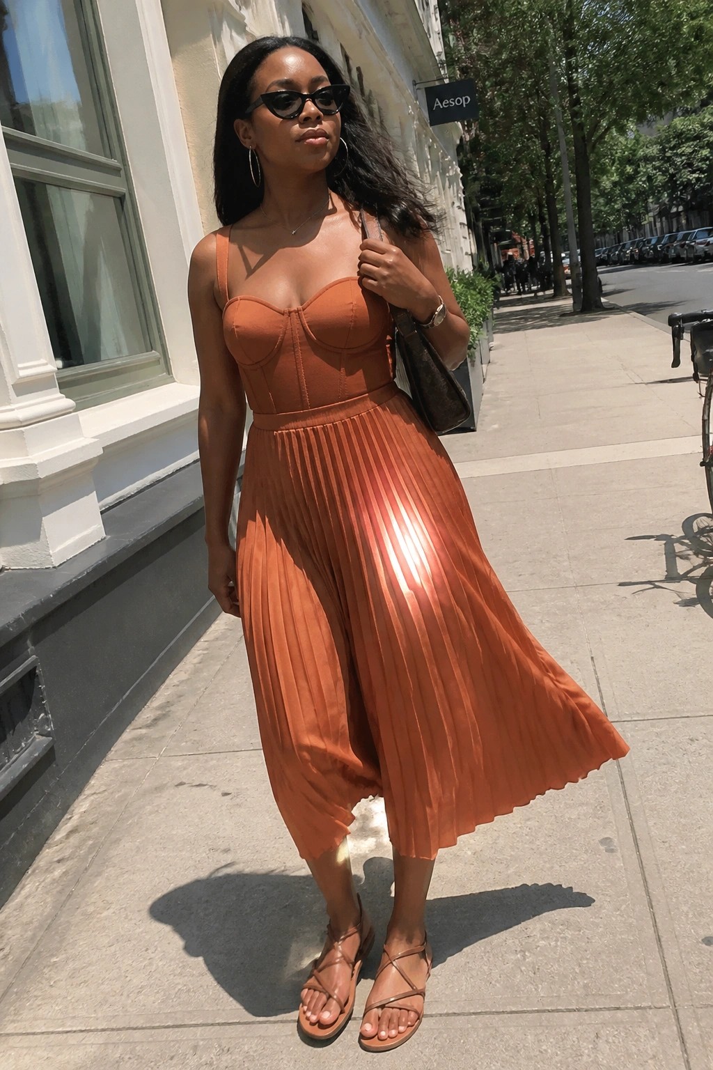 Woman in terracotta corset top and pleated midi dress on sunny city sidewalk