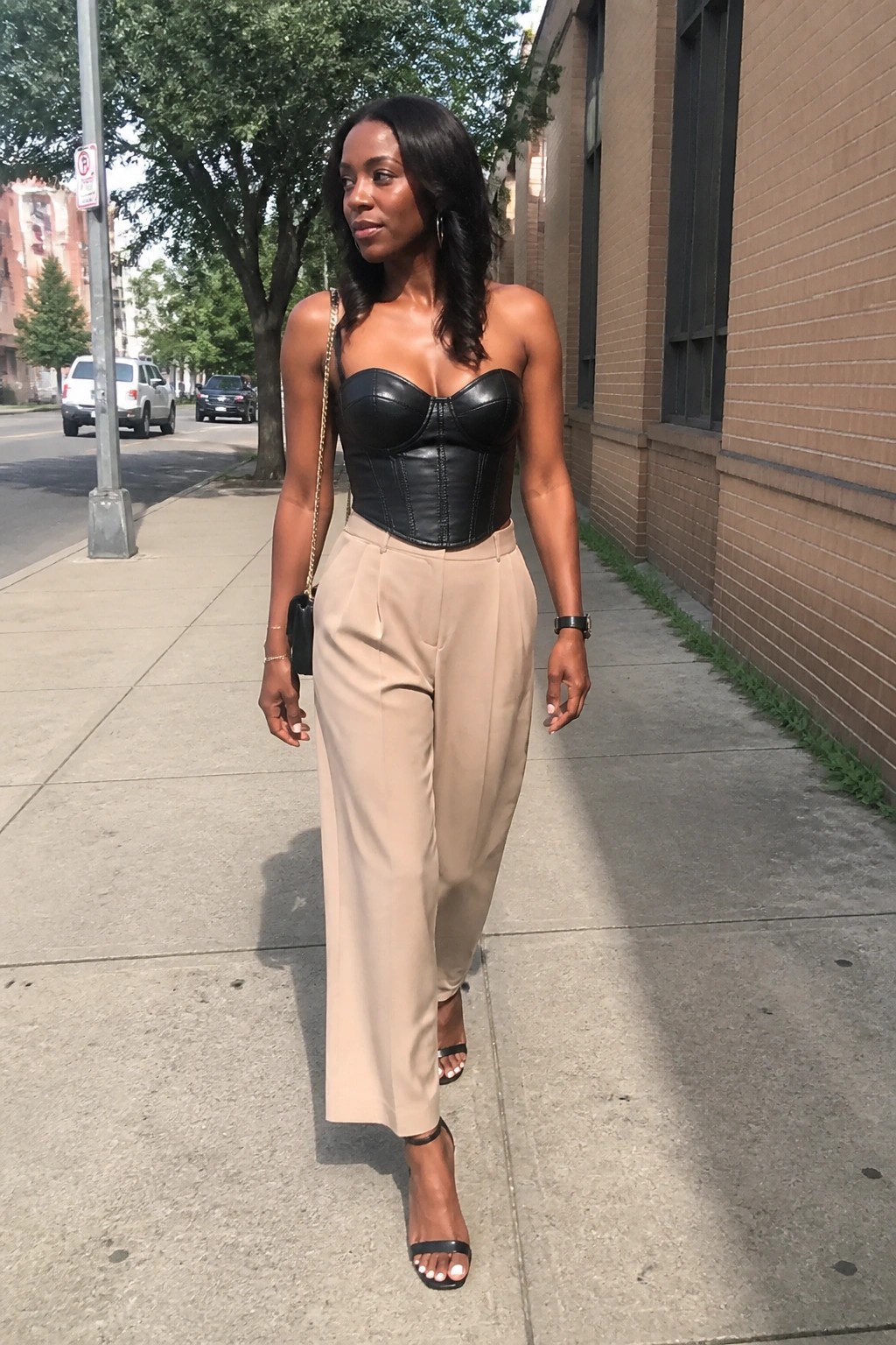 Stylish woman in black leather corset and beige wide-leg pants on sunny sidewalk.