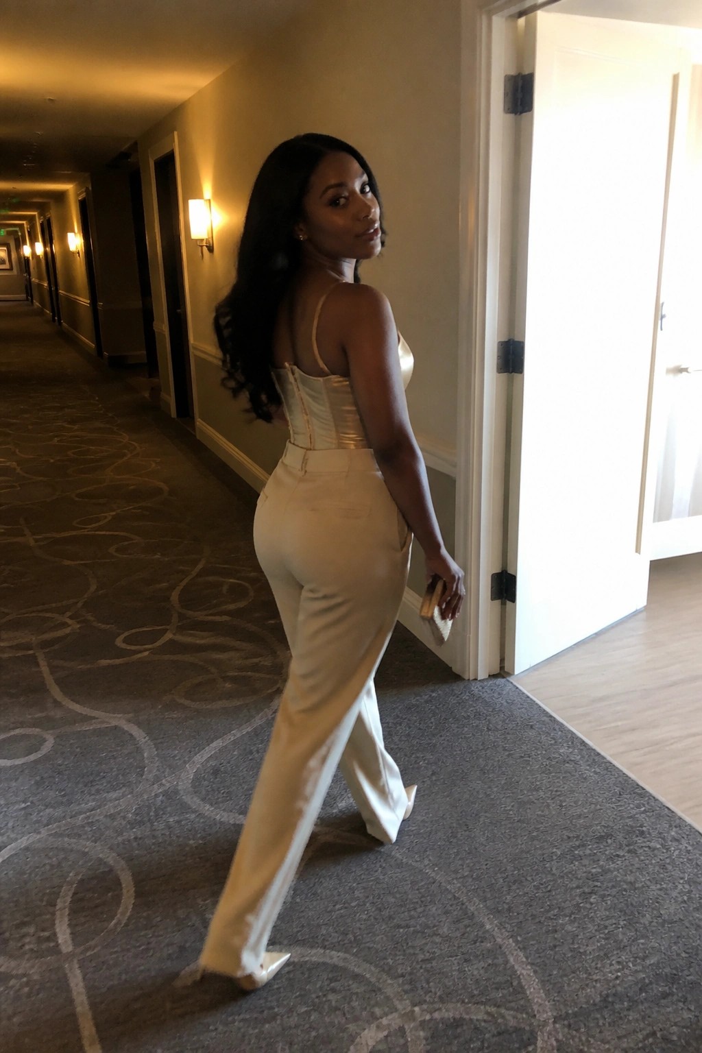 Woman in cream corset top and trousers glancing back in hotel hallway