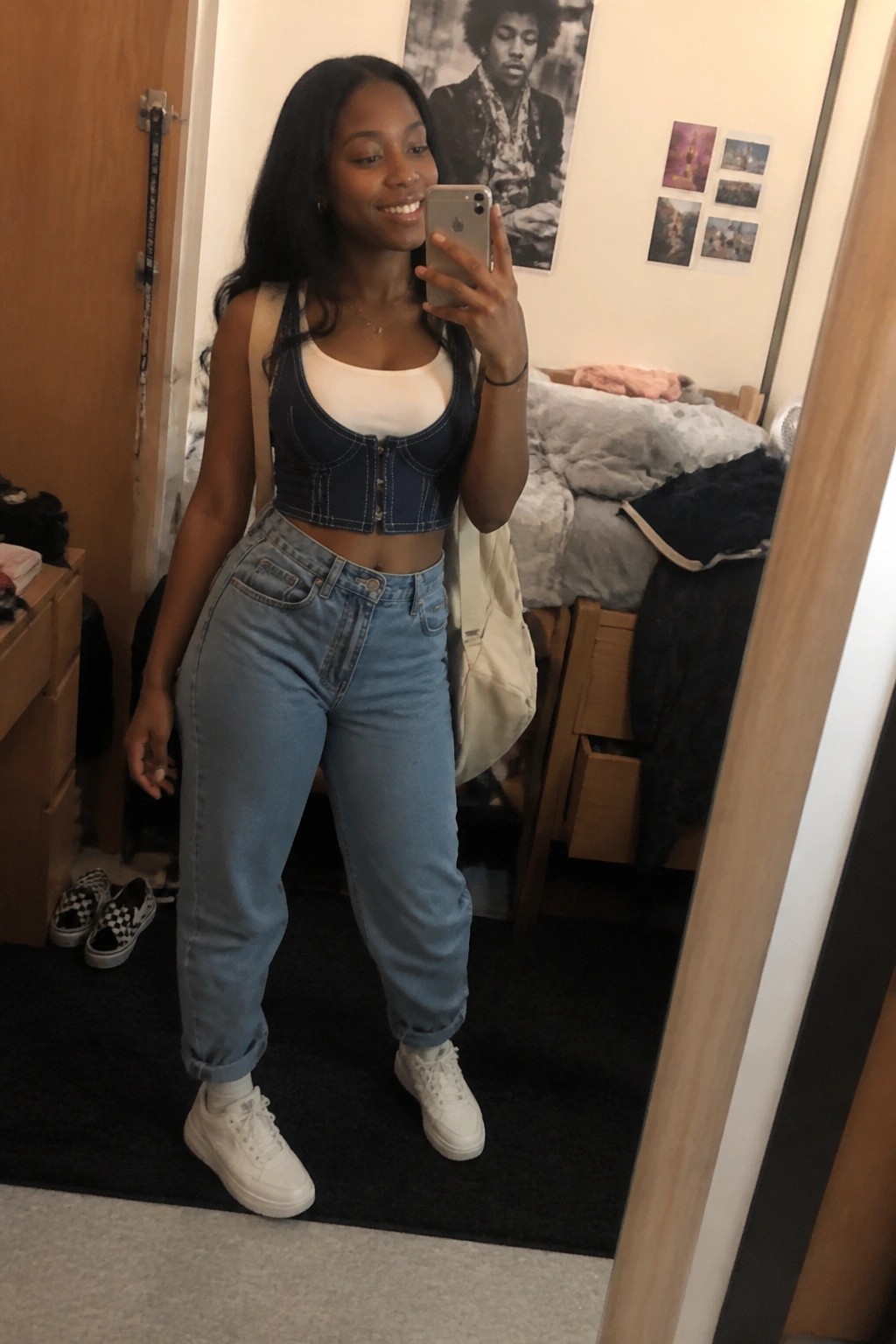 Woman wearing white tank under blue denim corset crop top, light wash mom jeans, white sneakers, and beige tote bag in a bedroom mirror selfie.