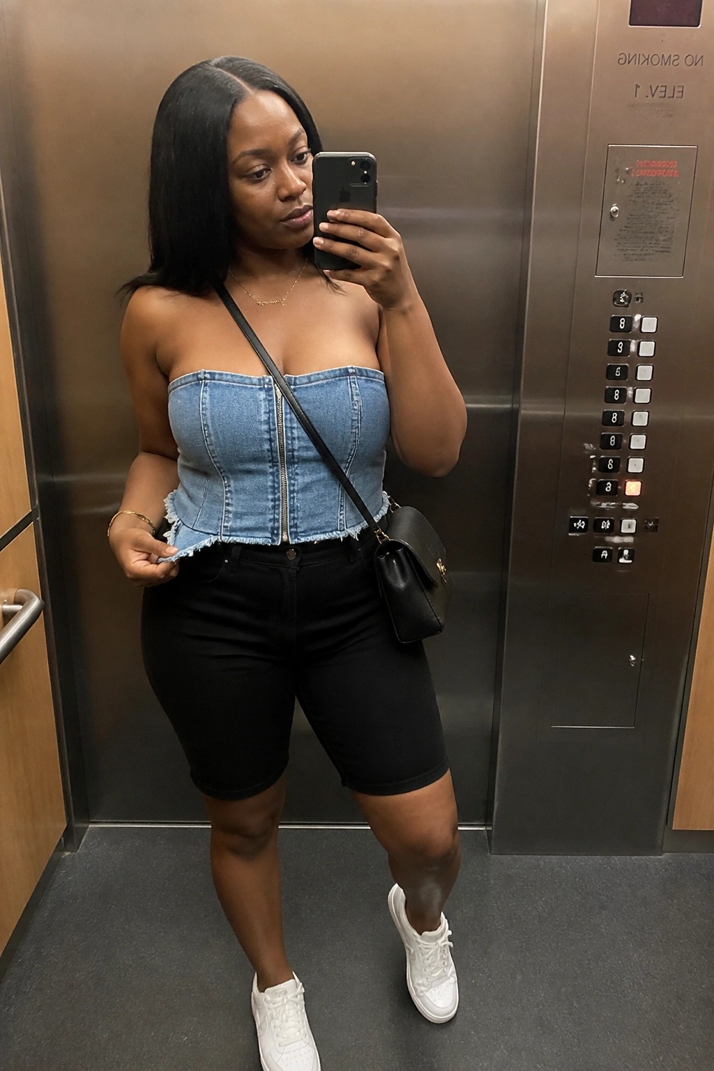 Woman in blue denim corset top and black shorts taking selfie in elevator