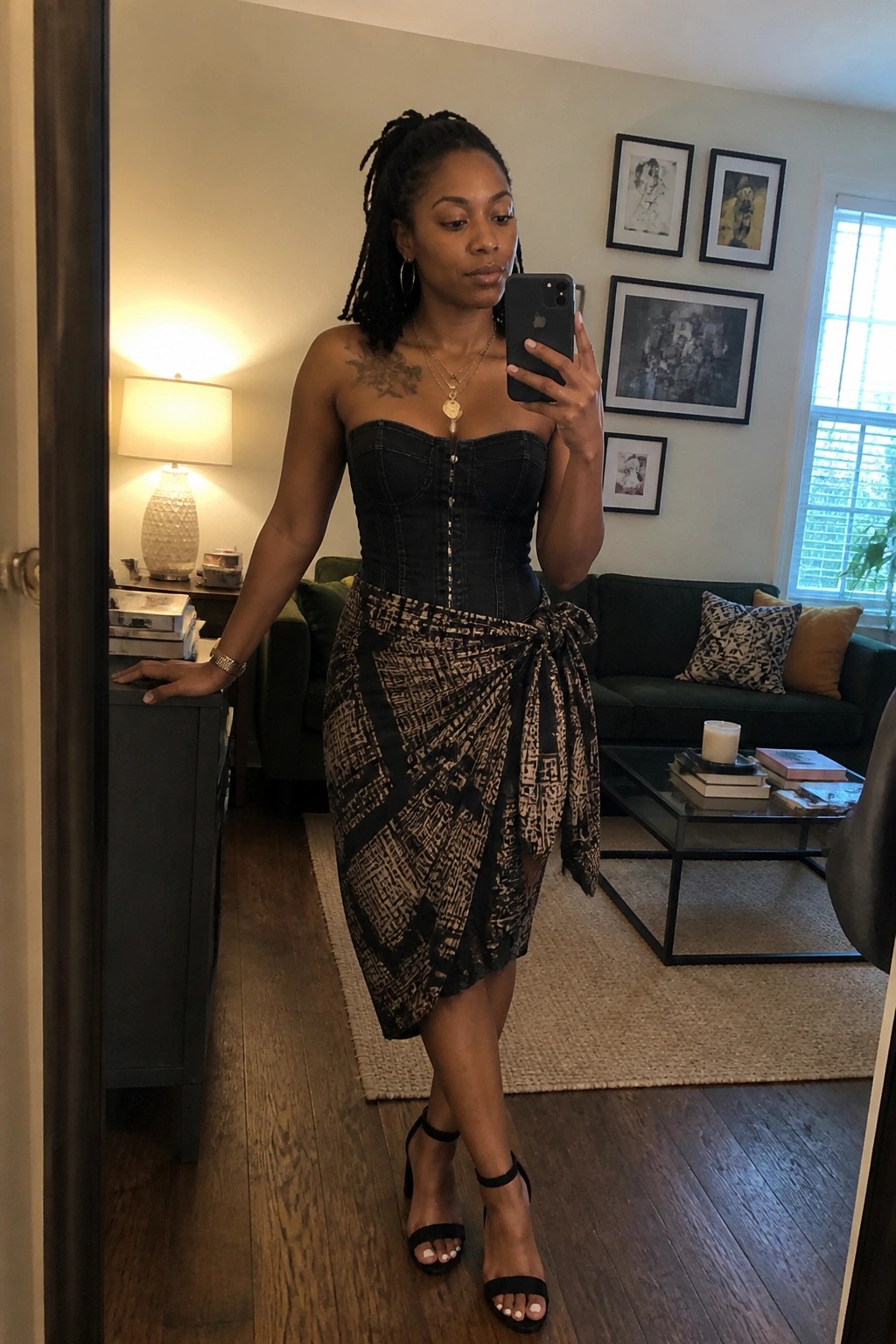 Woman in black denim corset and patterned wrap skirt taking mirror selfie in living room