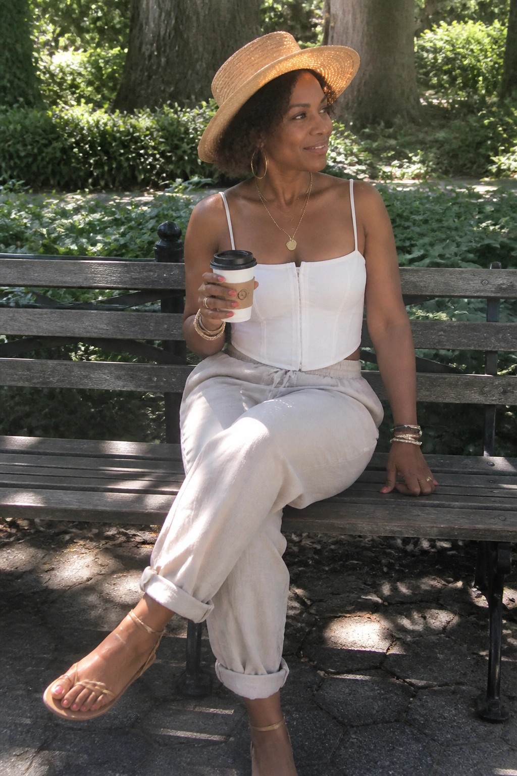 Woman in white corset top and beige pants holds coffee on park bench with straw hat