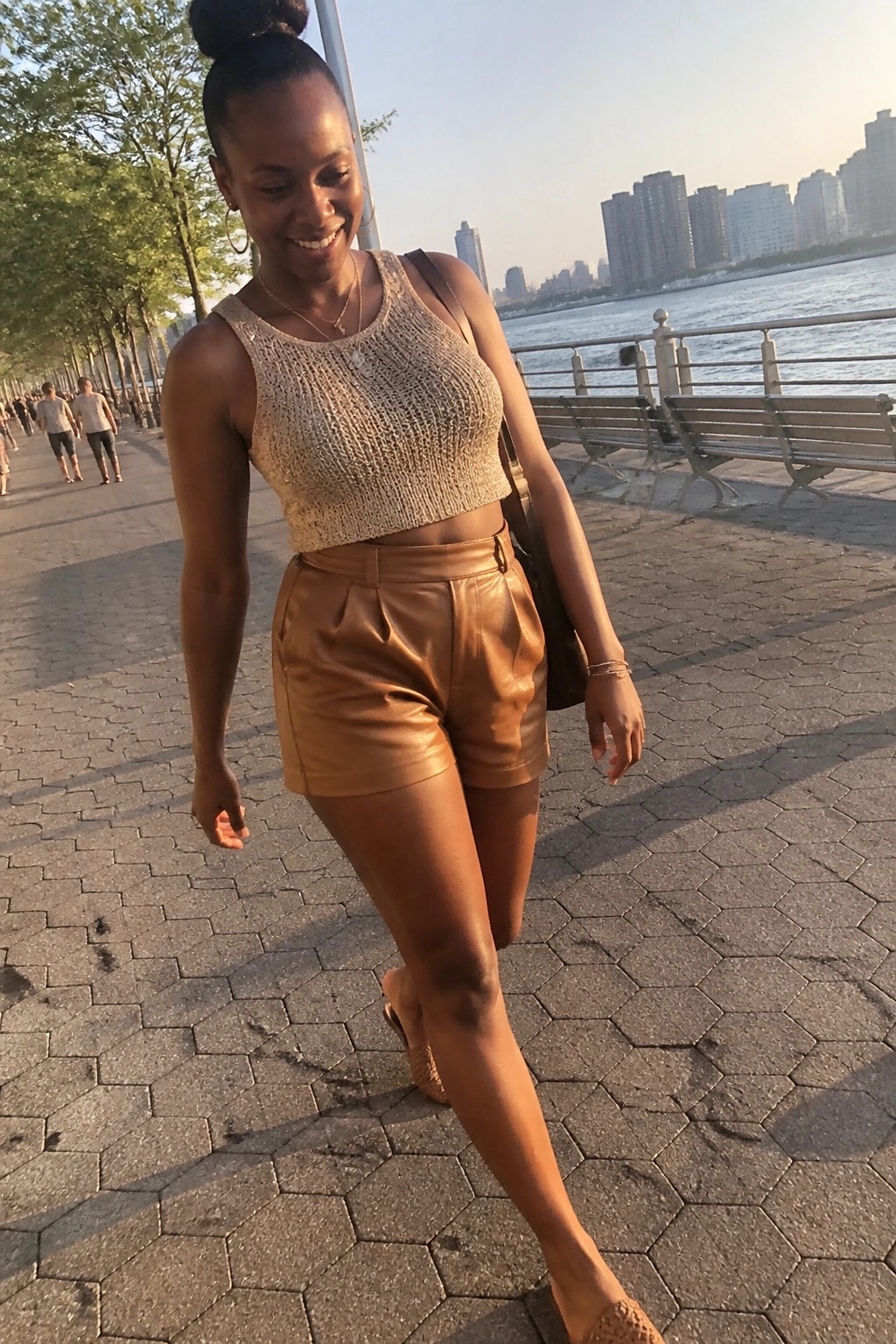 Smiling woman in beige knit crop top and brown leather shorts strolls along waterfront promenade at sunset.