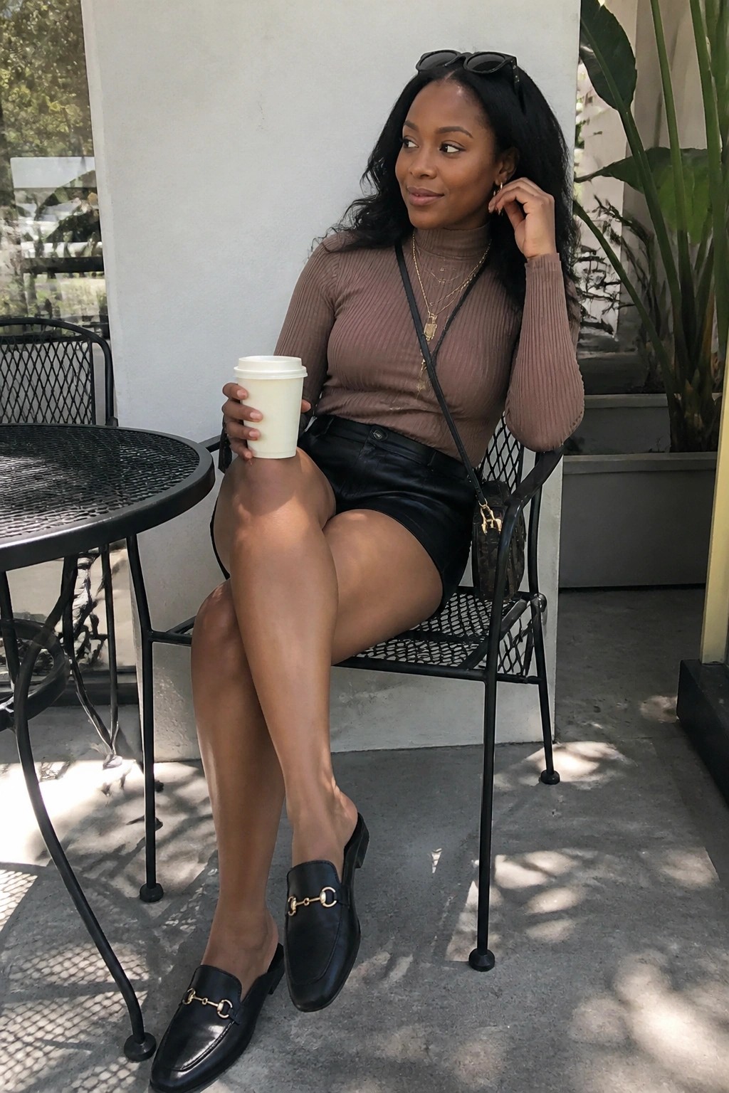 Woman in brown turtleneck and black leather shorts holds coffee outdoors