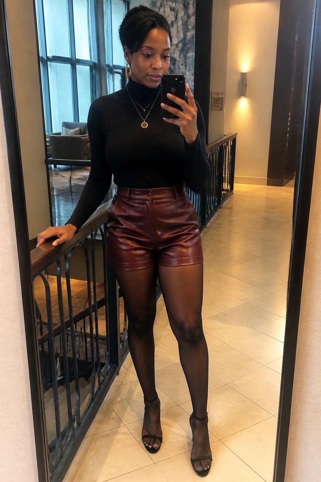 Woman in black turtleneck and maroon leather shorts taking mirror selfie in hallway