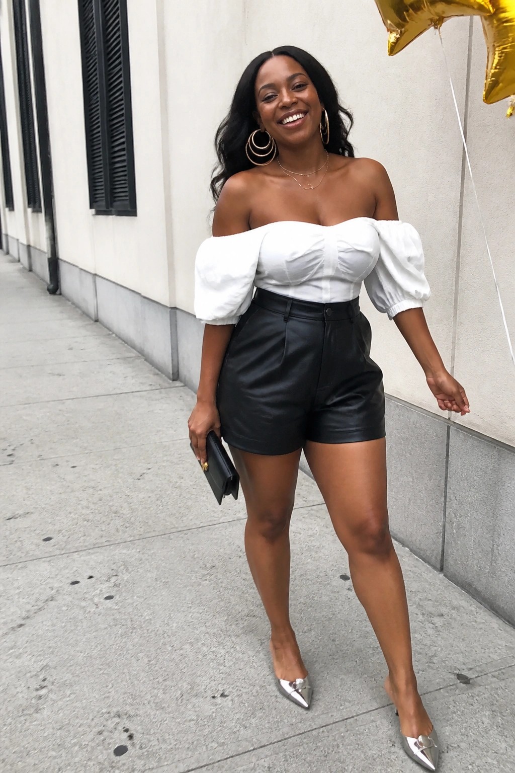 Woman in black leather shorts and white off-shoulder puff sleeve top with silver pointed heels and gold jewelry holding a black clutch.