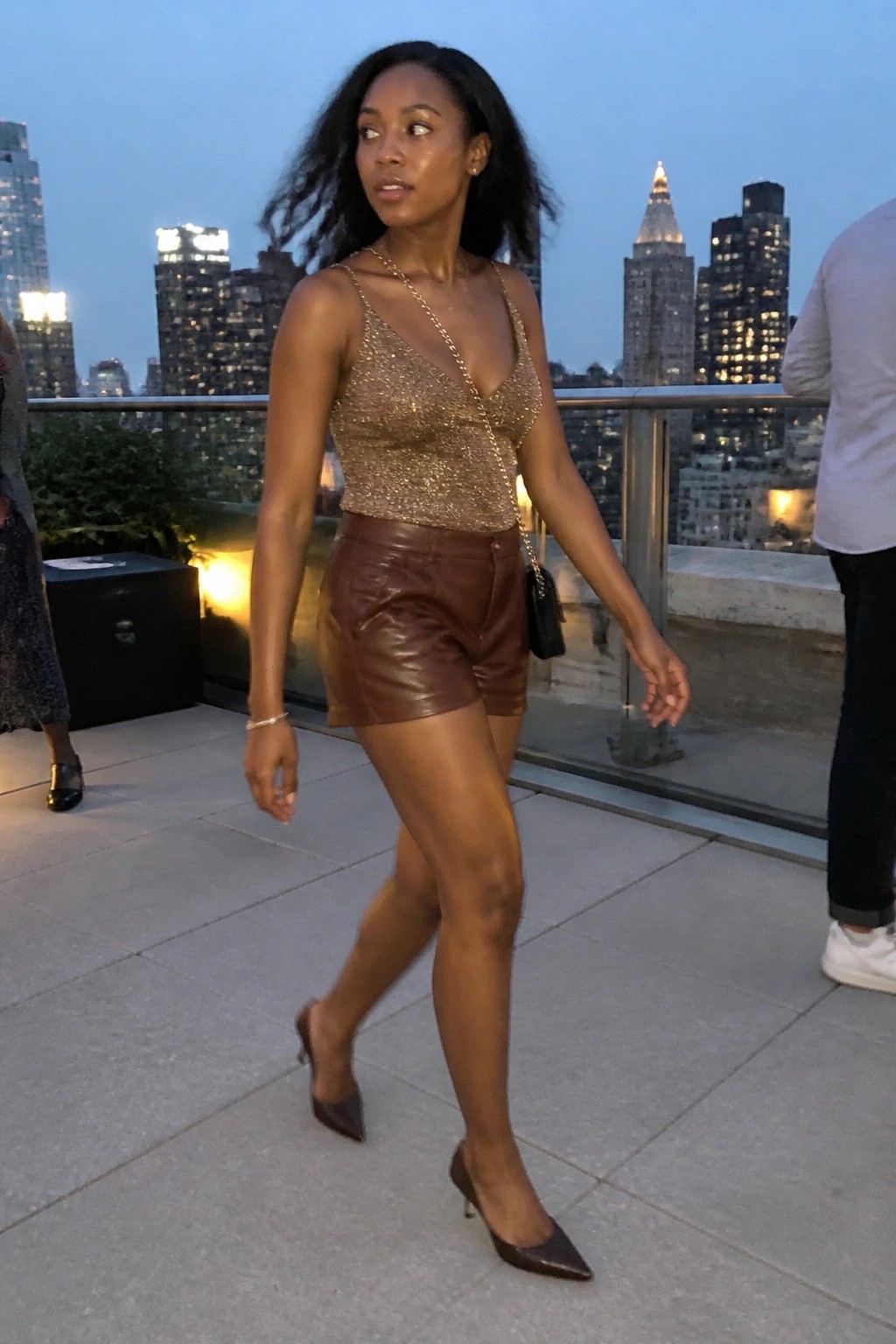 Woman in gold sequined top and brown leather shorts on rooftop at dusk