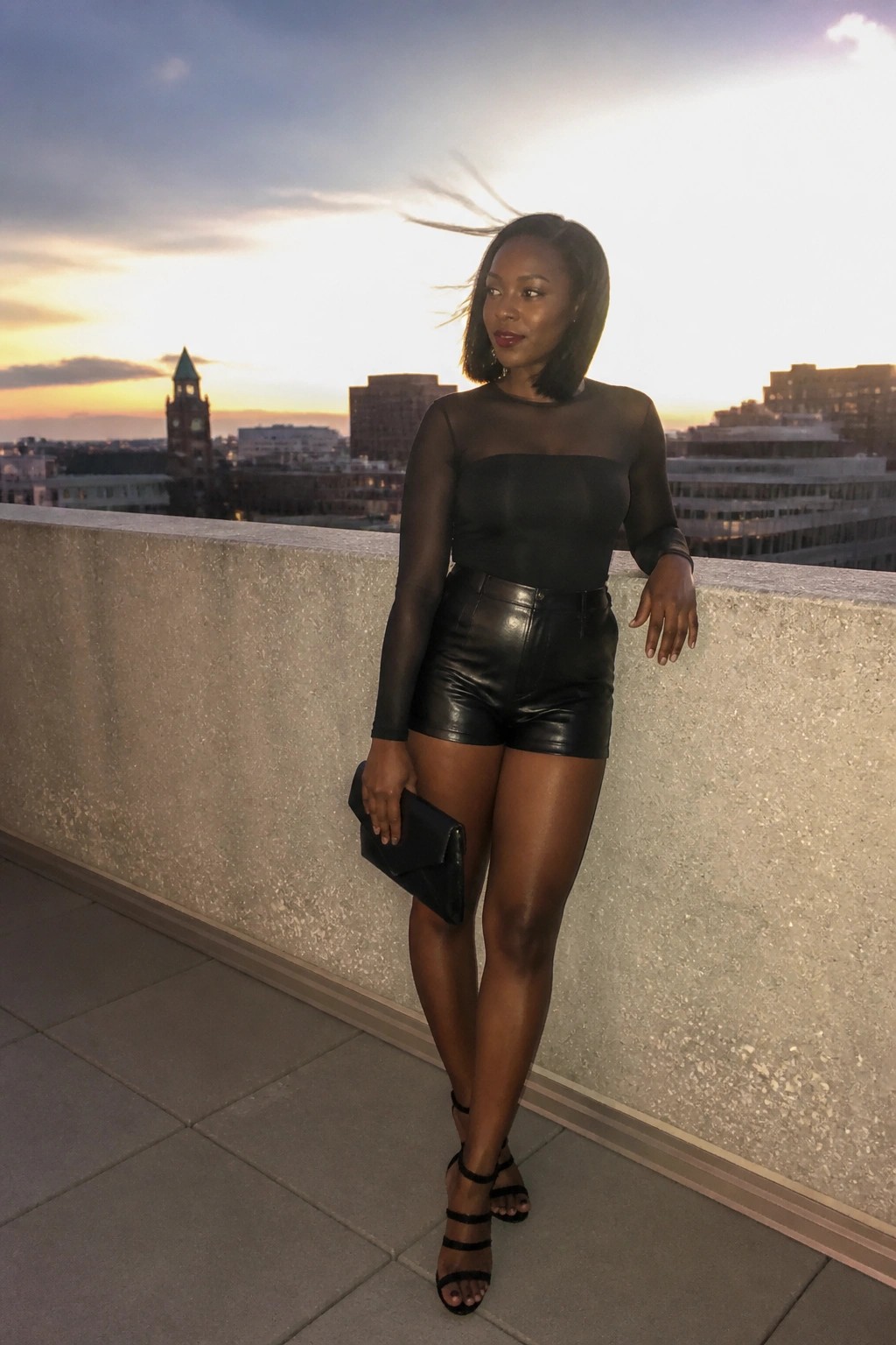 Woman in black sheer top and leather shorts posing on rooftop at sunset