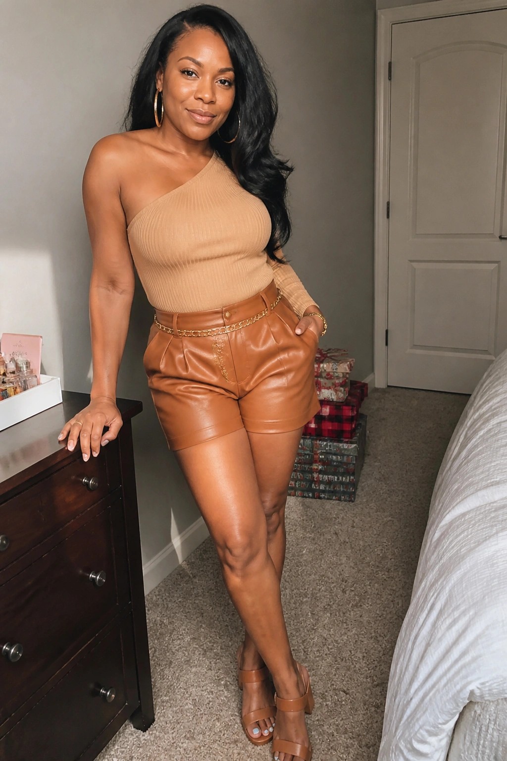 Woman in tan one-shoulder top and brown leather shorts posing by dresser in bedroom