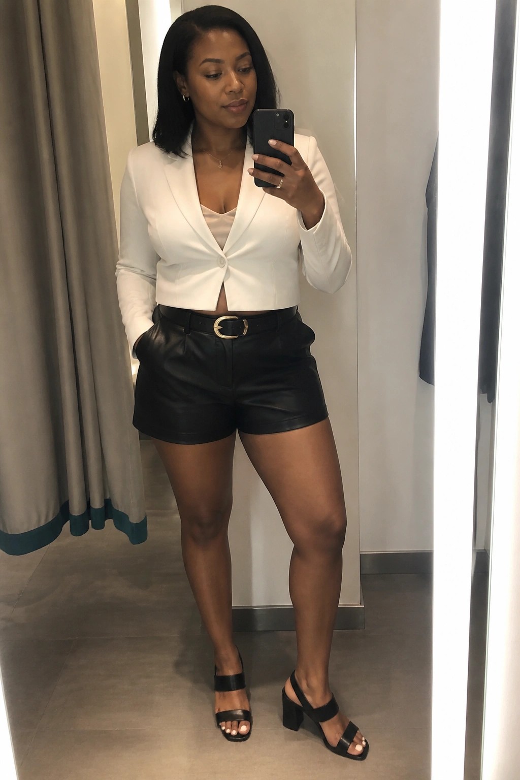 Woman in white cropped blazer and black leather shorts taking mirror selfie with phone
