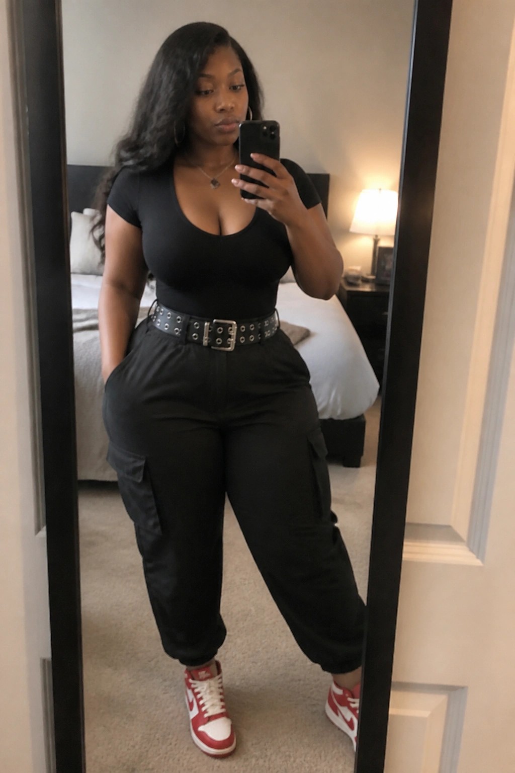 Woman in black top and cargo pants taking bedroom mirror selfie with red sneakers