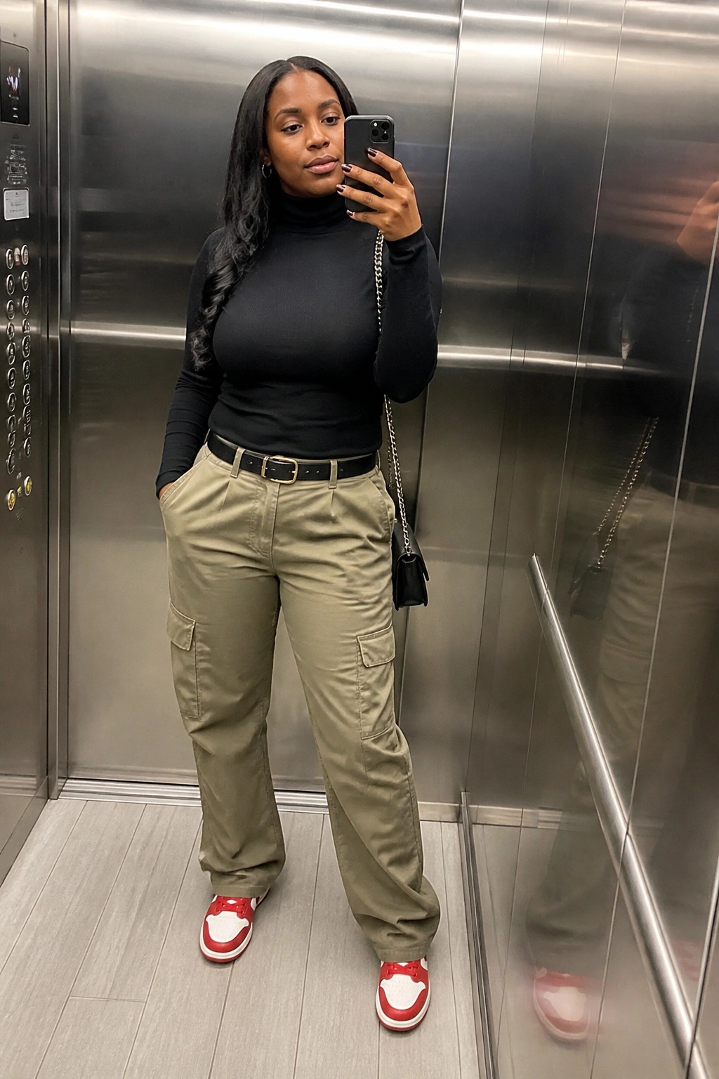 Woman in black turtleneck and olive cargo pants taking elevator selfie with phone.